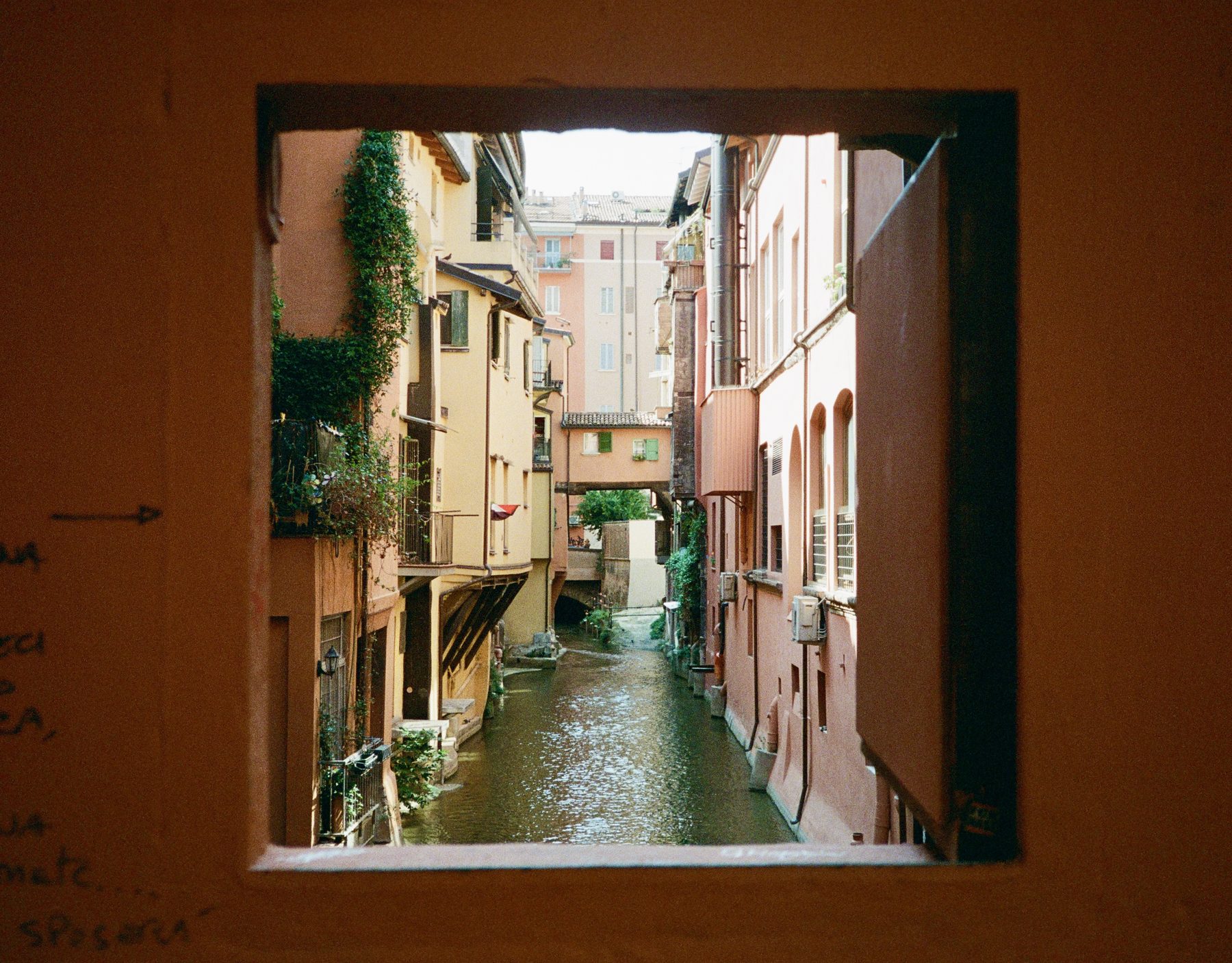 A photograph of a canal in Bologna, framed within an open window shutter.